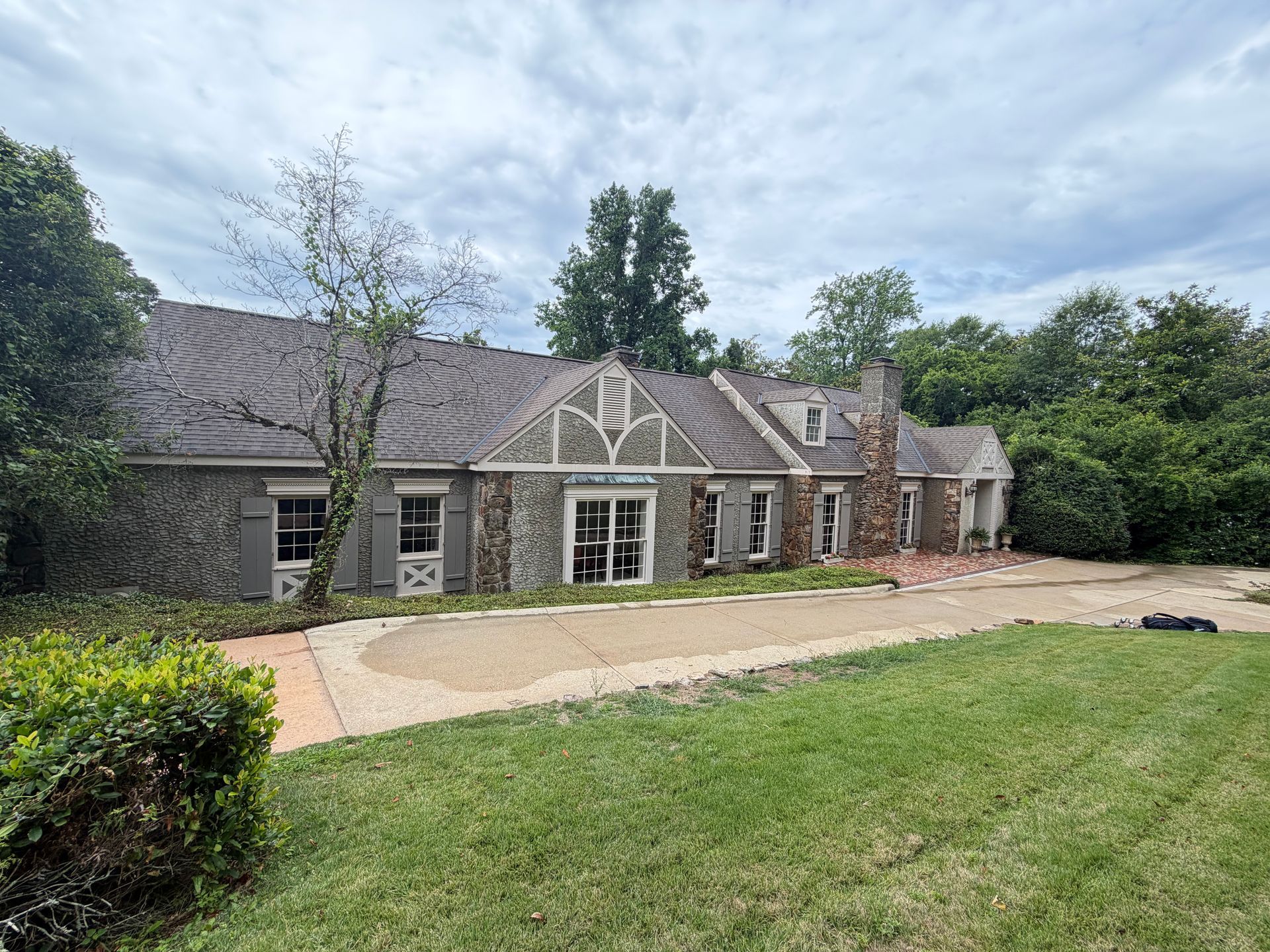 Stone house with a gray roof, front patio, and large green lawn under a cloudy sky