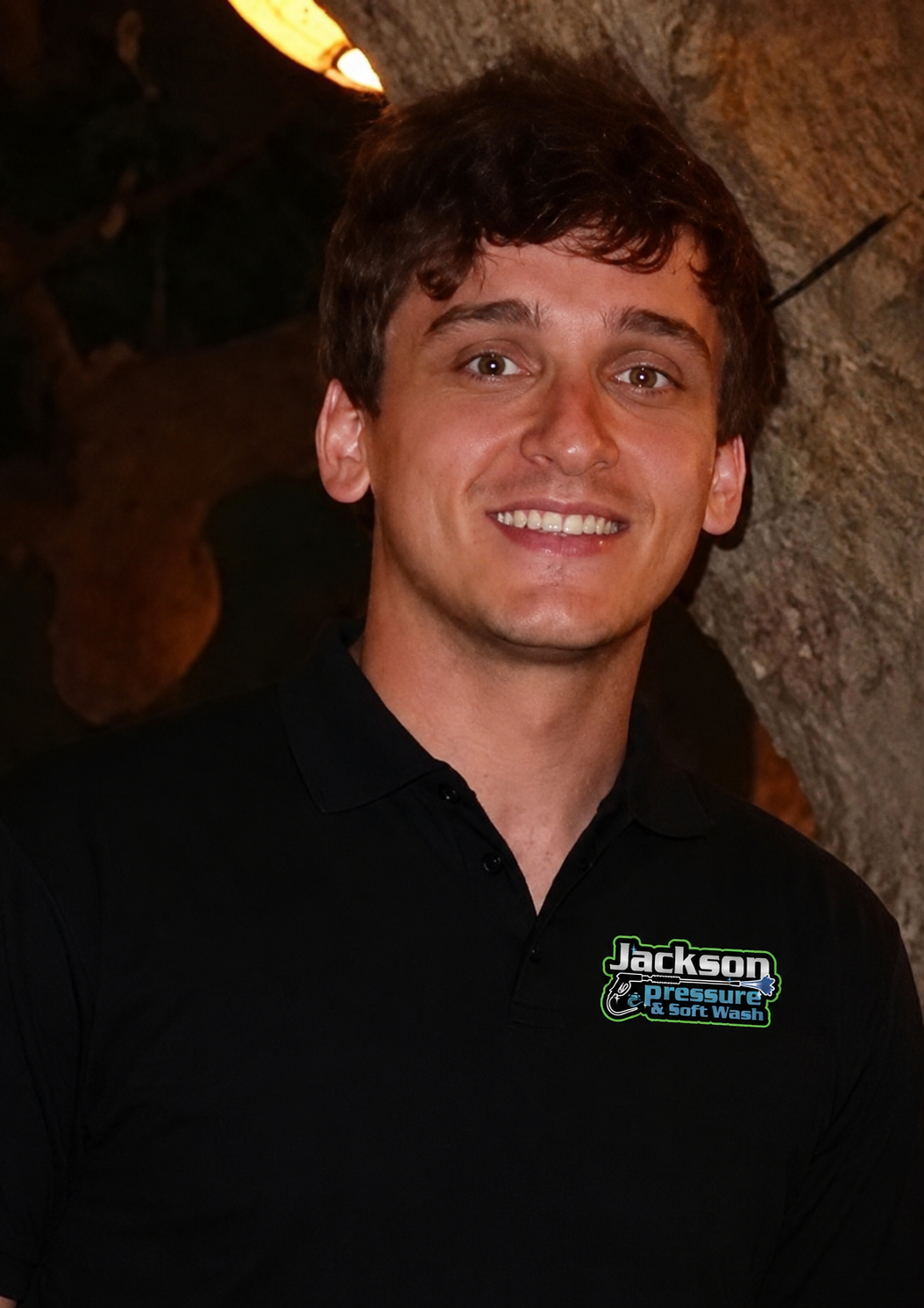 Smiling person in a black polo shirt standing by a tree outdoors at night