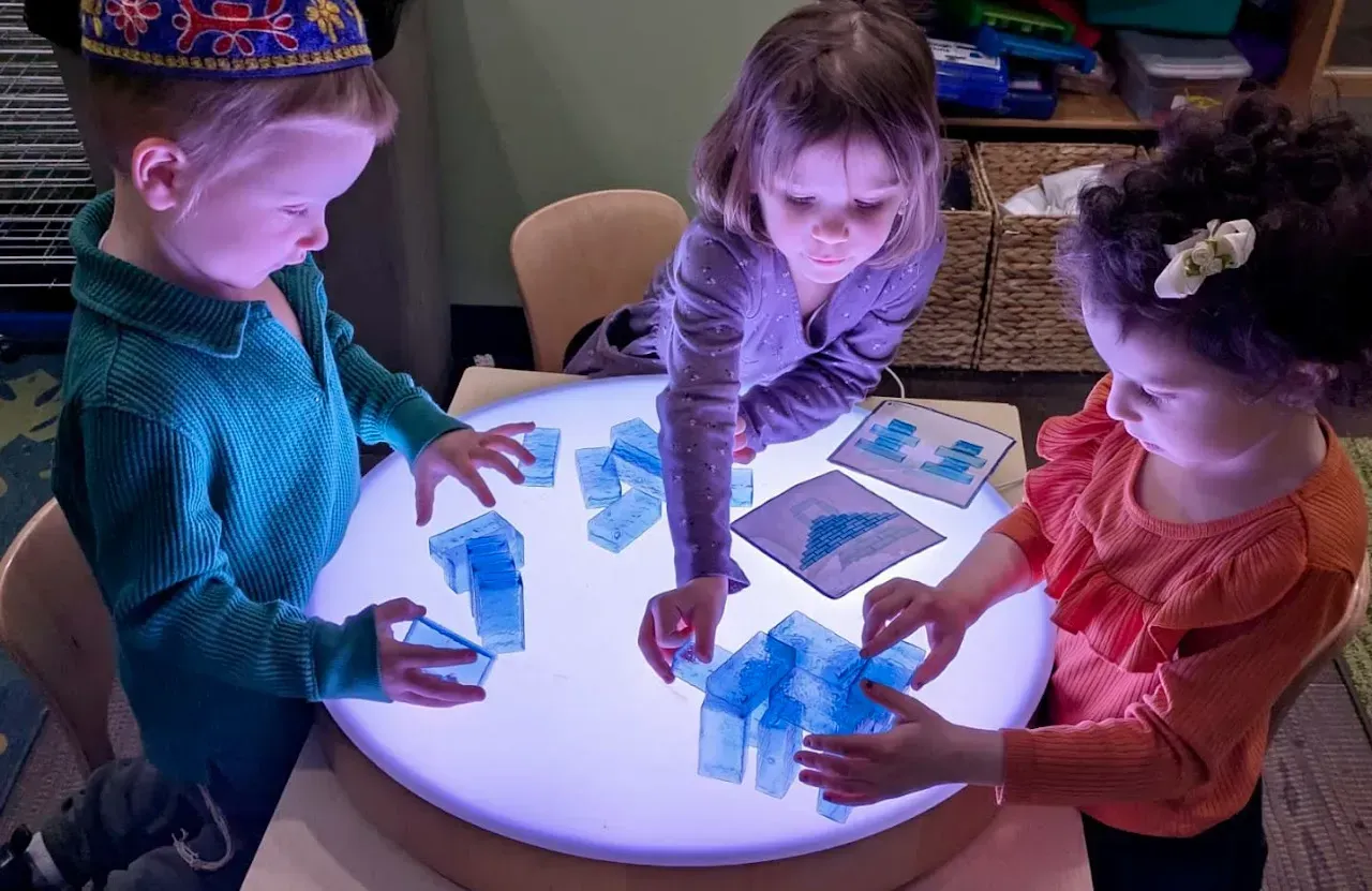 Children play with colorful blocks around a glowing round table in a classroom.