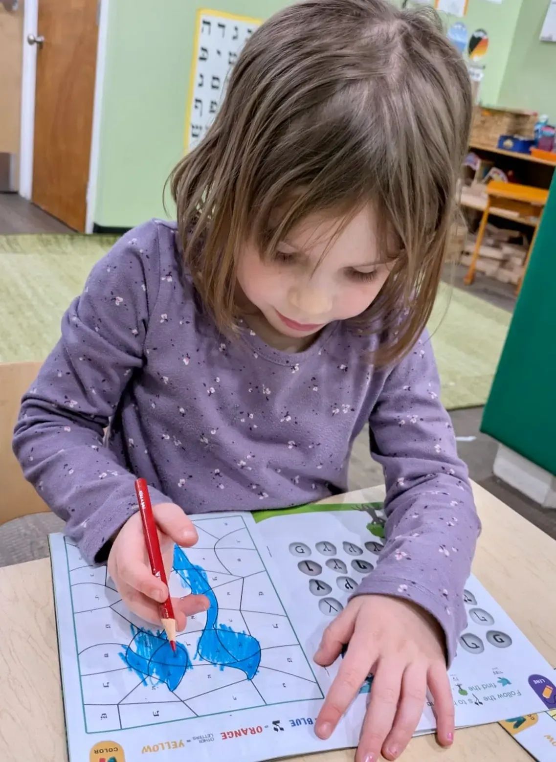 Child coloring on a worksheet at a classroom table with a red marker