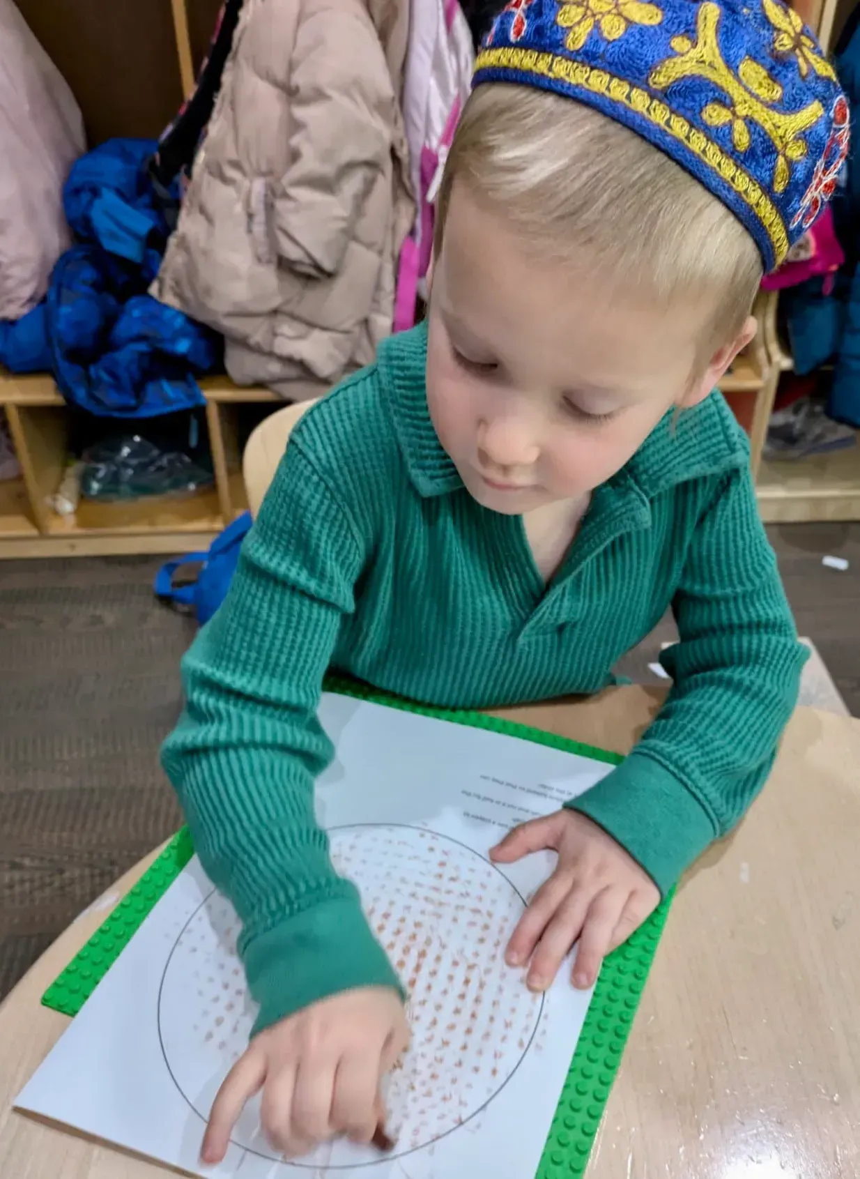 Child in a green sweater tracing a dotted circle on paper at a classroom table.