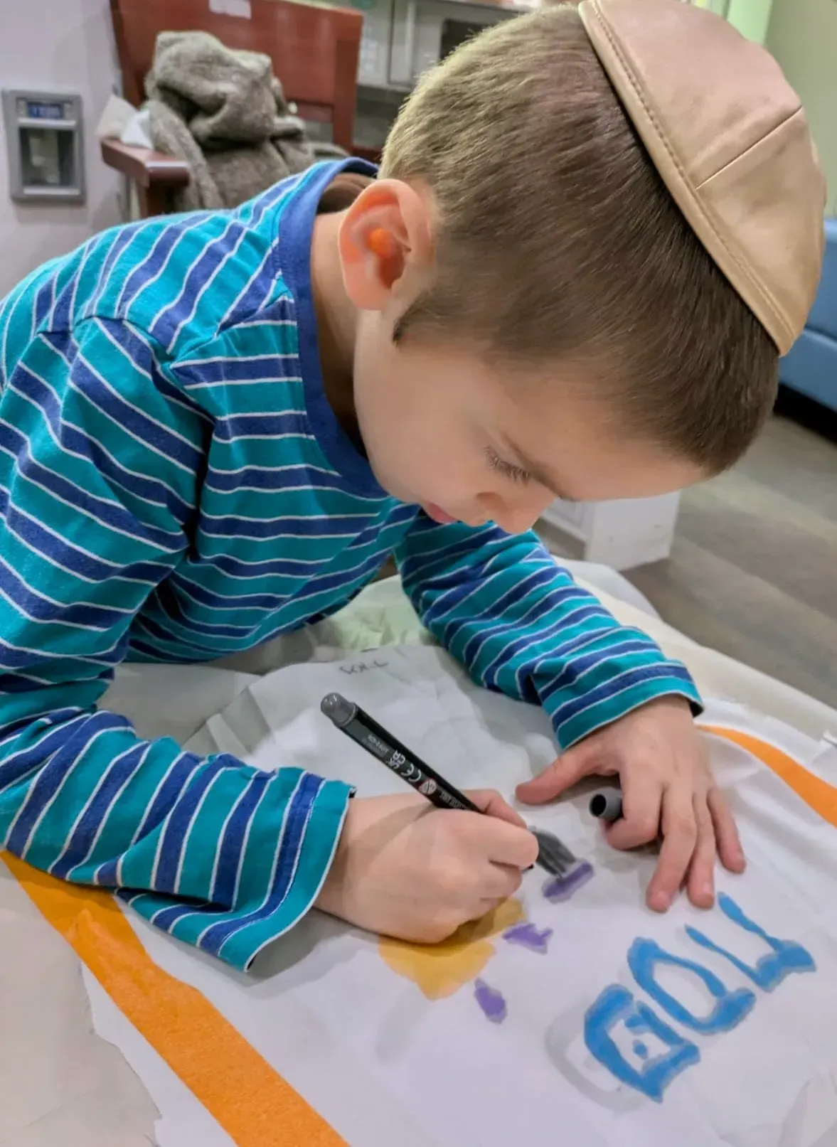 Child in a kippah draws on paper with a marker at a table.