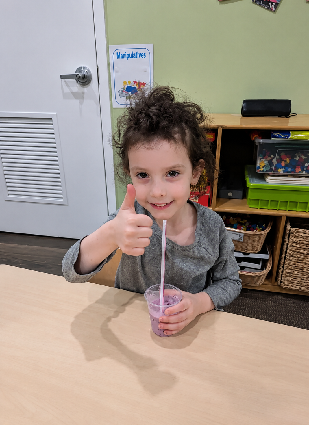 Child at a table giving a thumbs-up while stirring a drink with a straw in a classroom.