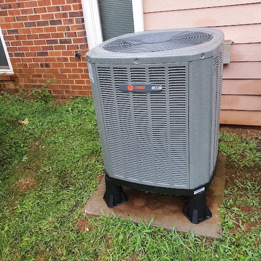 A gray air conditioner is sitting in the grass in front of a brick house.