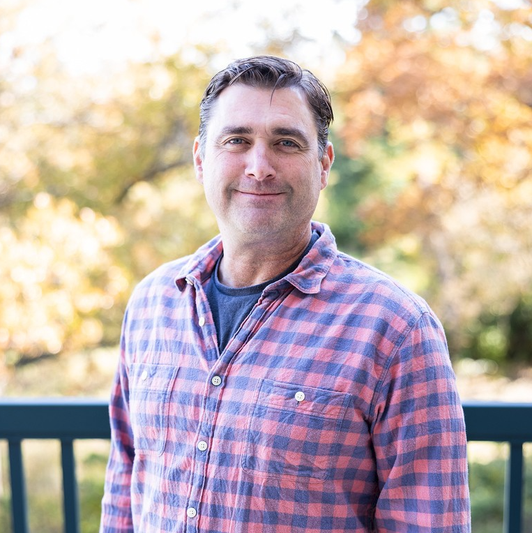 Man in red plaid shirt smiling, standing outside with fall foliage in background.