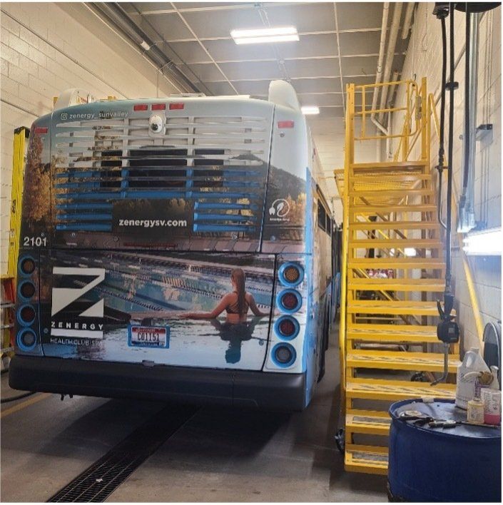Back of a bus with a water scene wrap, parked in a maintenance bay, next to yellow stairs.