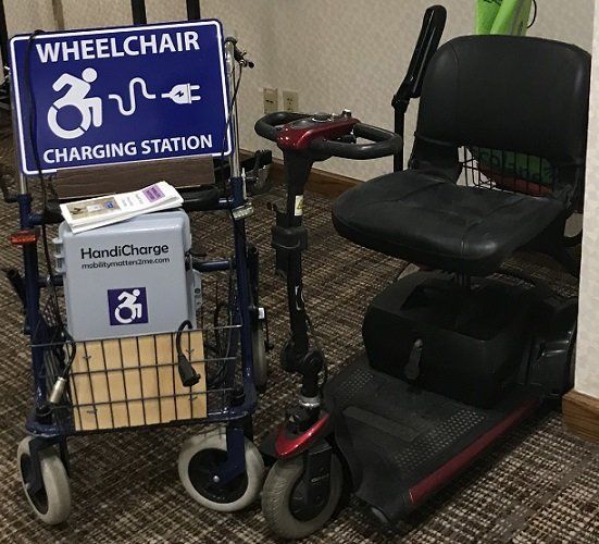 Wheelchair charging station next to mobility scooter and walker. Blue sign with wheelchair symbol and plug icon.