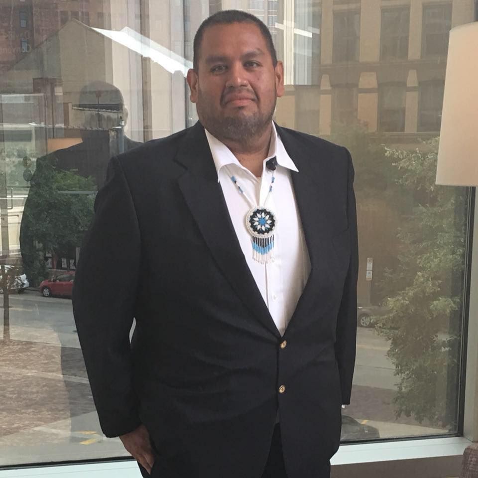 Man in suit and beaded necklace stands near a window.