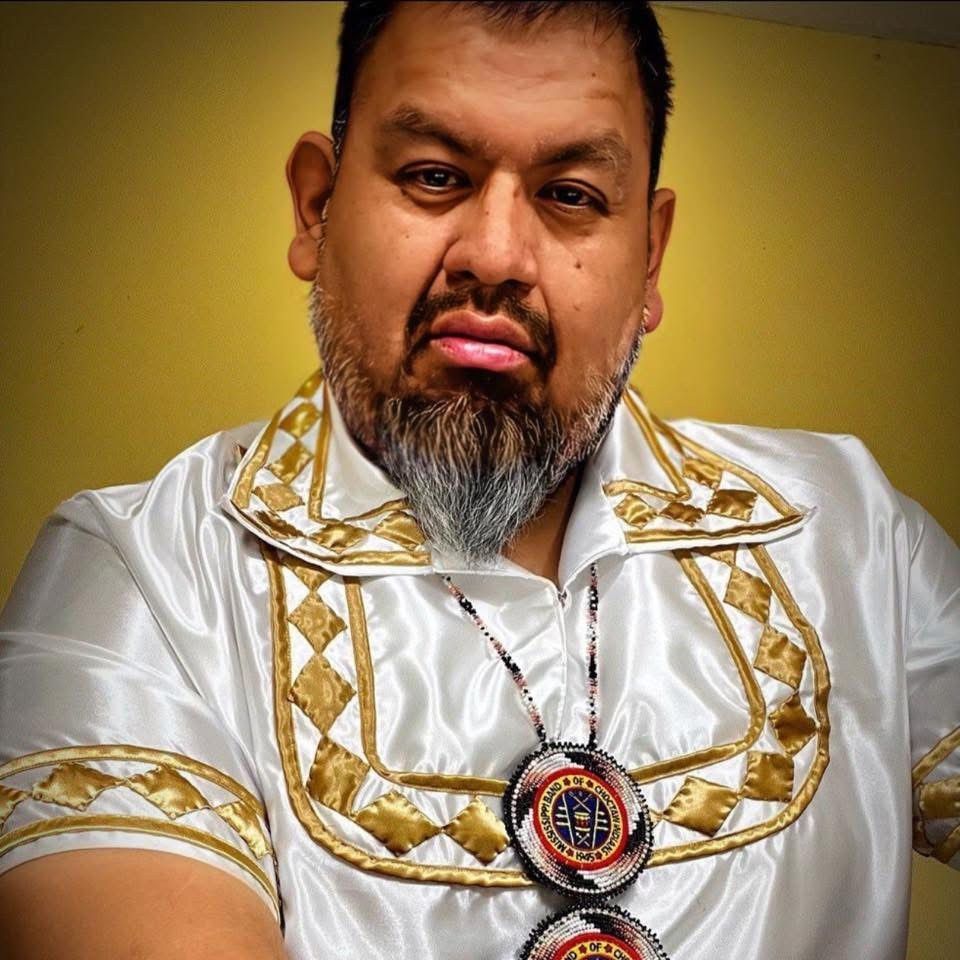 Man with dark hair and beard wearing a white shirt with gold trim, beaded necklace.