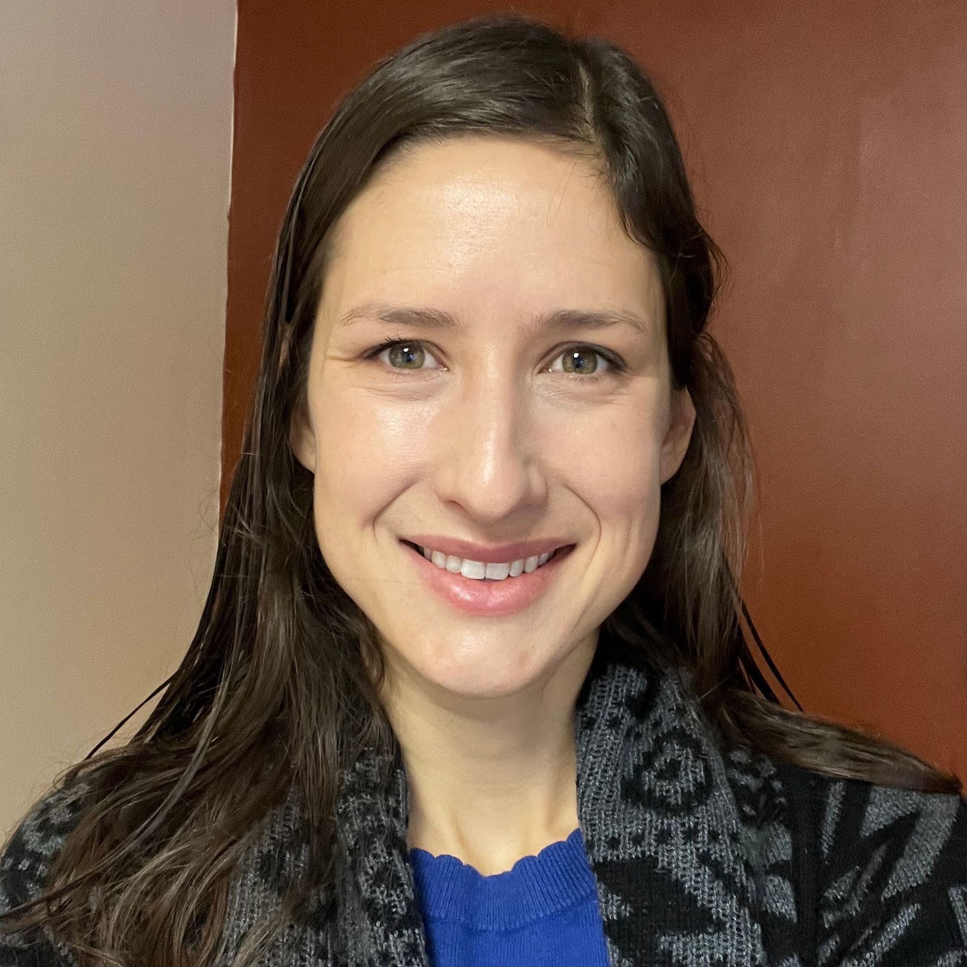 Woman with long brown hair smiles at the camera; wearing a blue shirt and patterned sweater.
