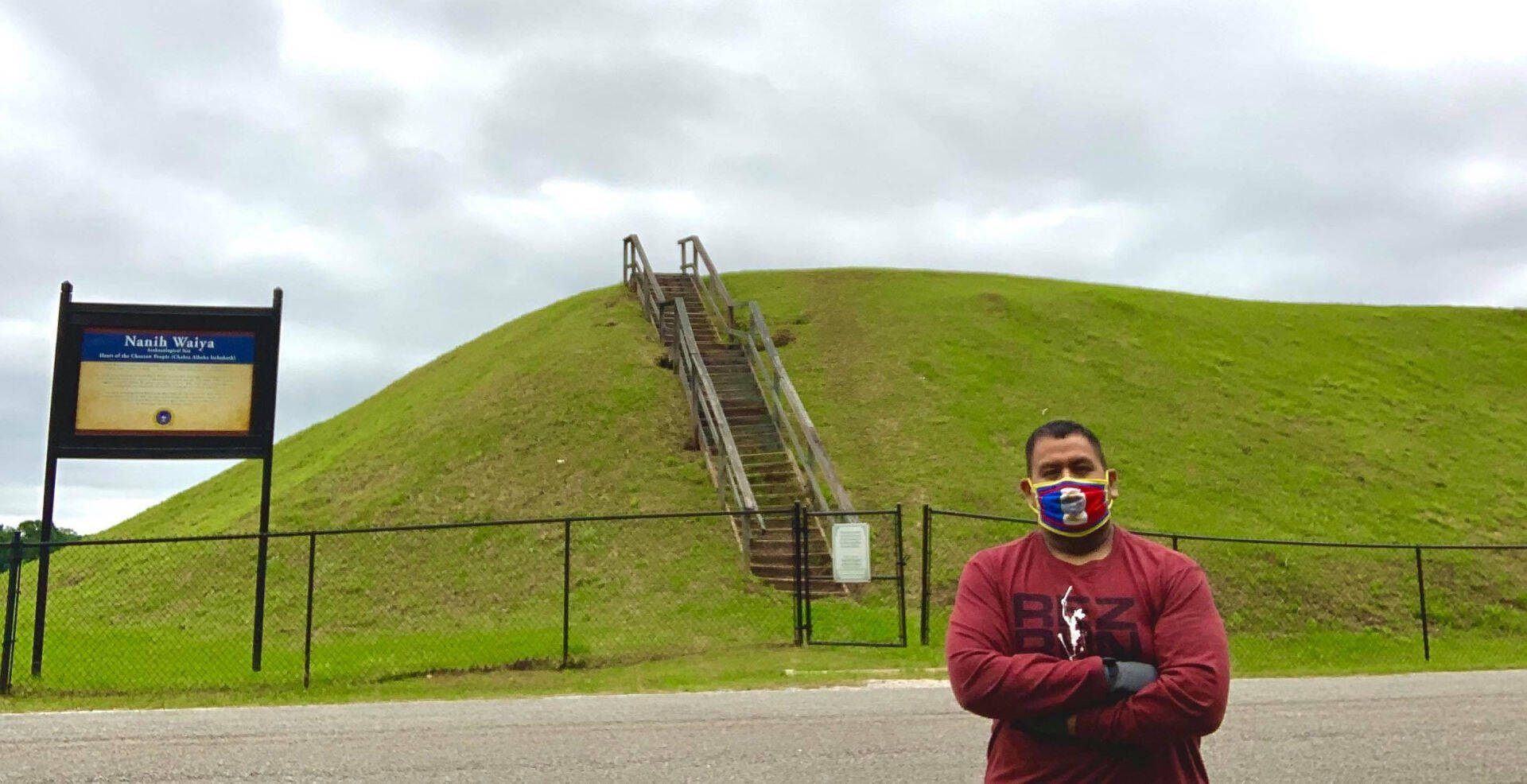 Man wearing a mask stands in front of a large mound with wooden stairs. A sign and fence are also present. Cloudy sky.