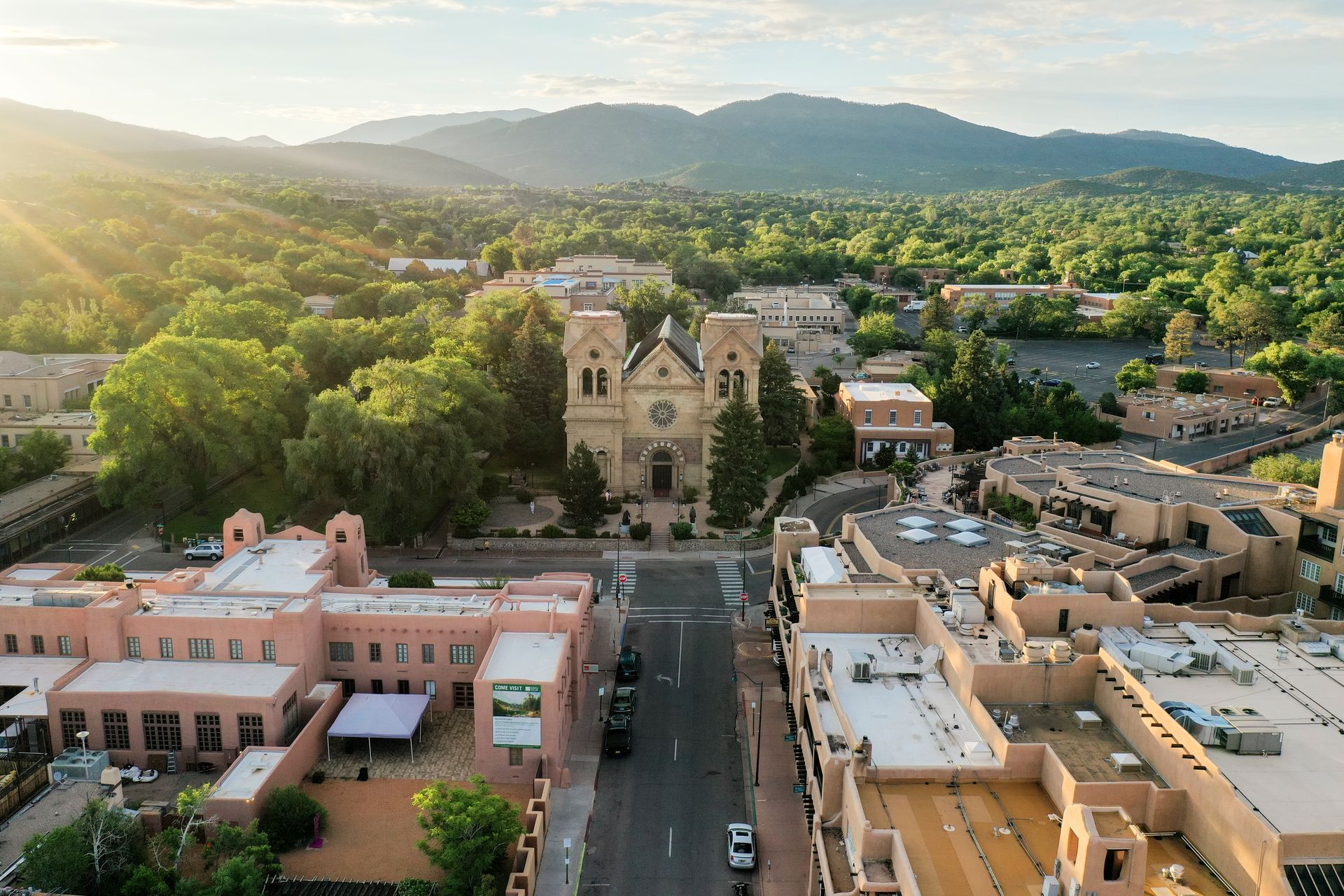 Aerial view of Santa Fe, New Mexico, with a large cathedral at the center, surrounded by buildings and lush greenery.