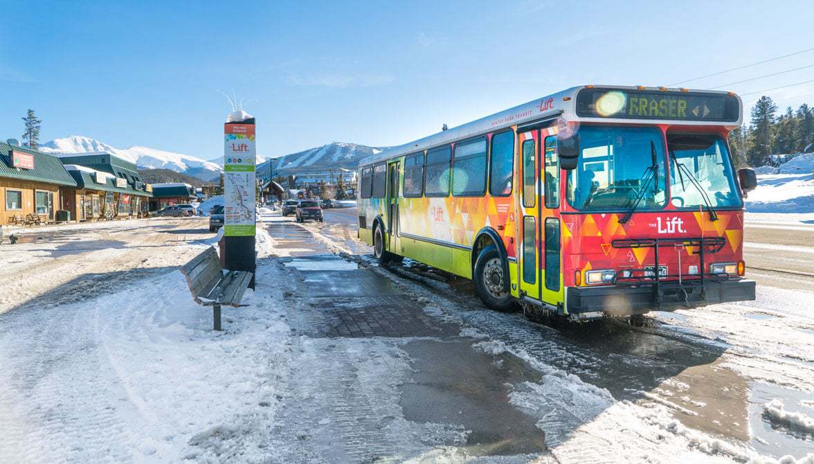 Bus at a snowy stop with mountains in the background; colorful artwork on bus side.