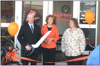 Three people cut a ribbon at a bicycle shop opening. Orange balloons, a bike, and shop sign visible.