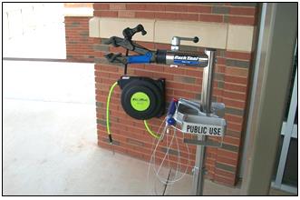Air pump station for public use, against brick wall; features air hose, pressure gauge, and hanging tools.