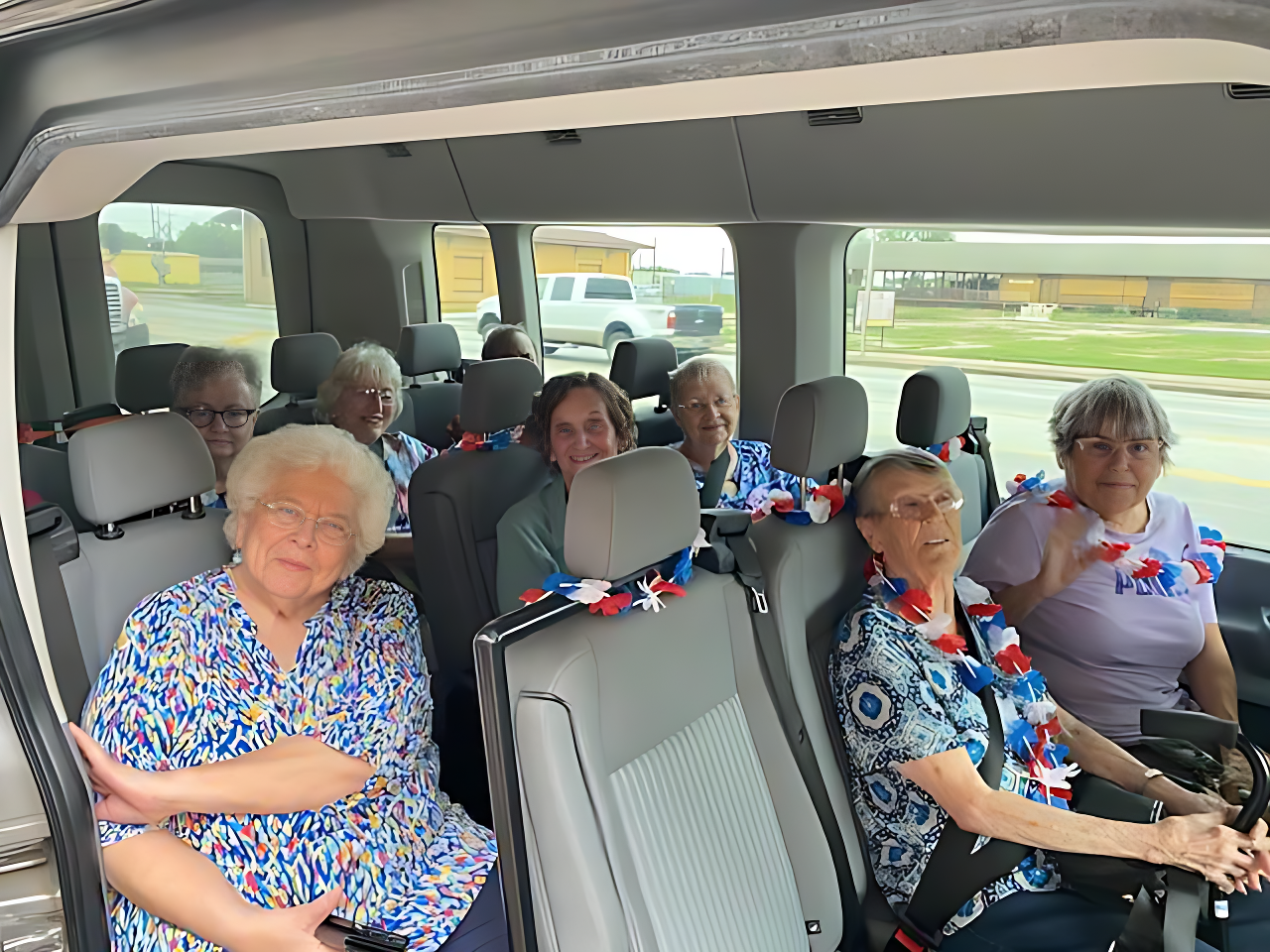 Seniors inside a van, smiling, wearing patriotic garlands. Grey seats, sunny outdoors in background.