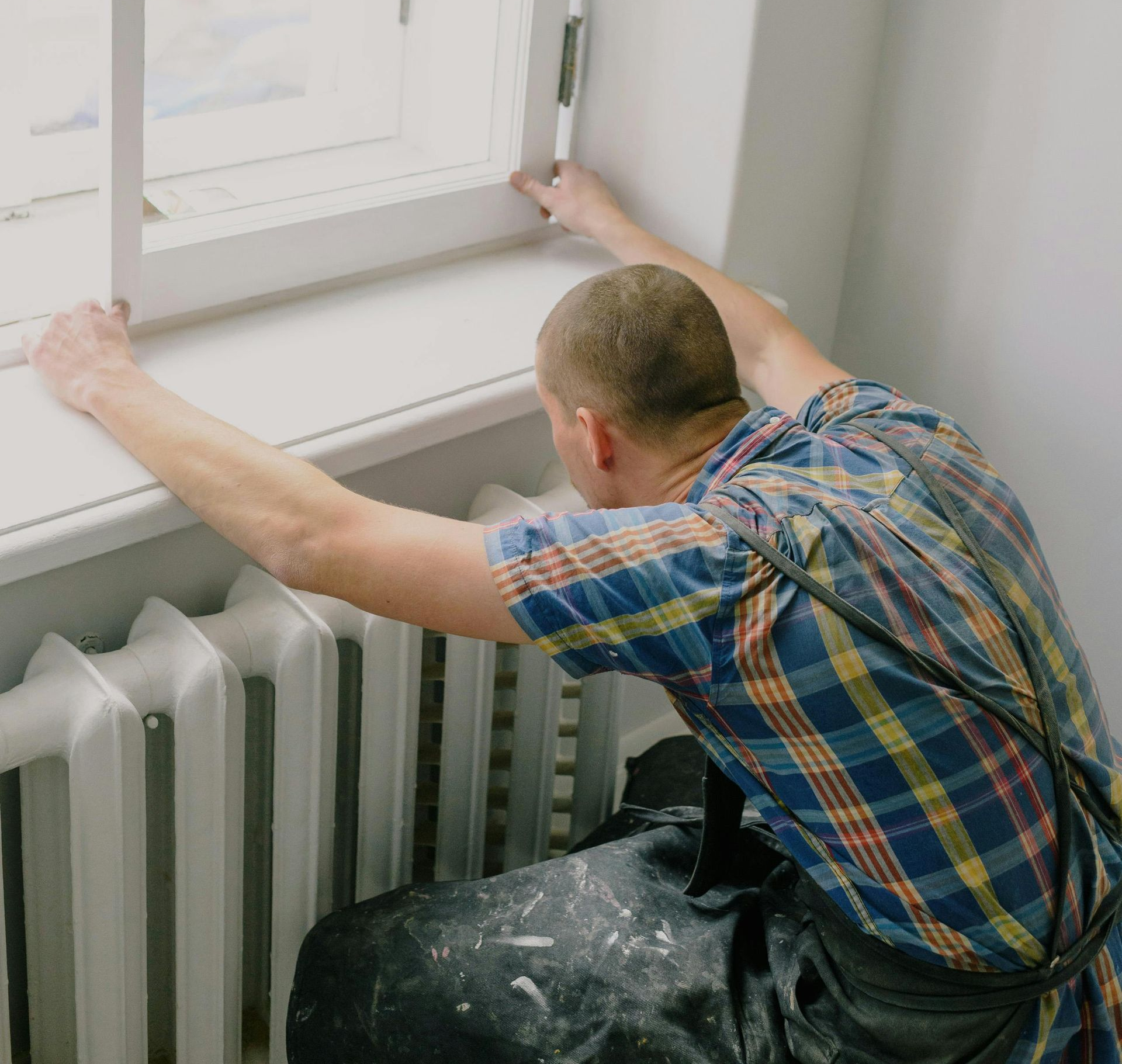 Man leans over a window frame, likely inspecting it. He wears a plaid shirt and overalls, standing in a room with a radiator.
