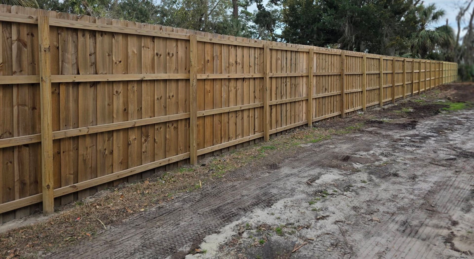 Wooden privacy fence along a muddy dirt path beside a tree-lined area