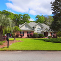 Suburban brick home with white gables, green lawn, palm trees, and a clear blue sky