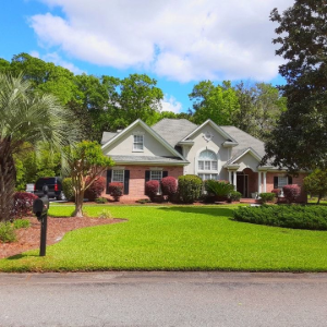 Suburban brick home with white gables, green lawn, palm trees, and a clear blue sky