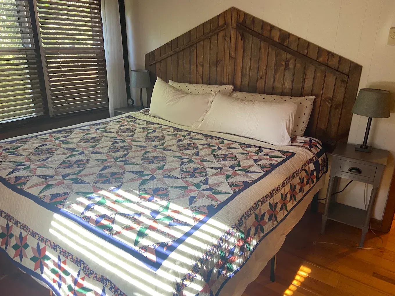 Bedroom with a wooden headboard, patterned quilt, white pillows, and a bedside lamp by a window.