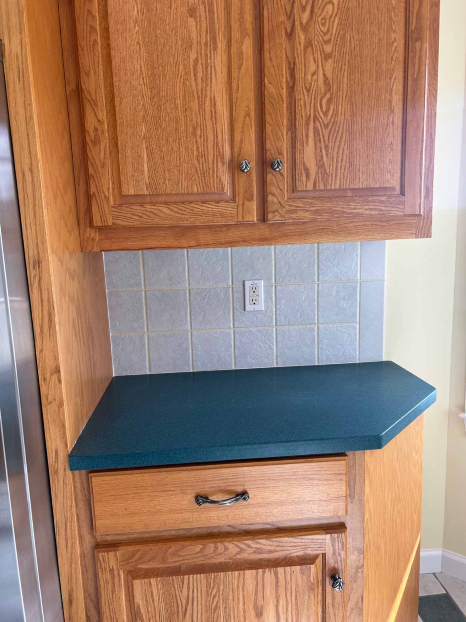 Wooden kitchen cabinets above a teal countertop with a tiled backsplash and electrical outlet