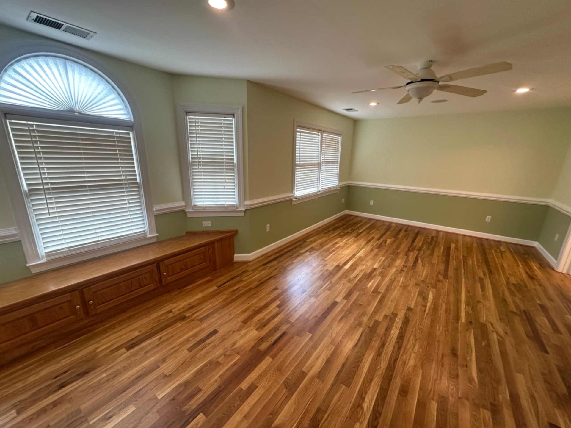 Empty living room with hardwood floors, green walls, large windows, and a ceiling fan.