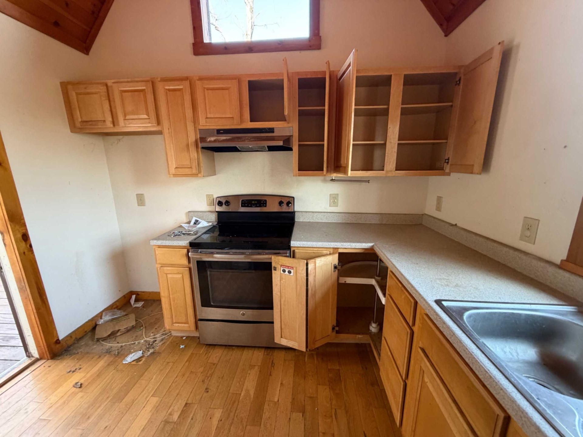 Empty kitchen with wooden cabinets, stainless stove, sink, and open cabinet doors under a small window.
