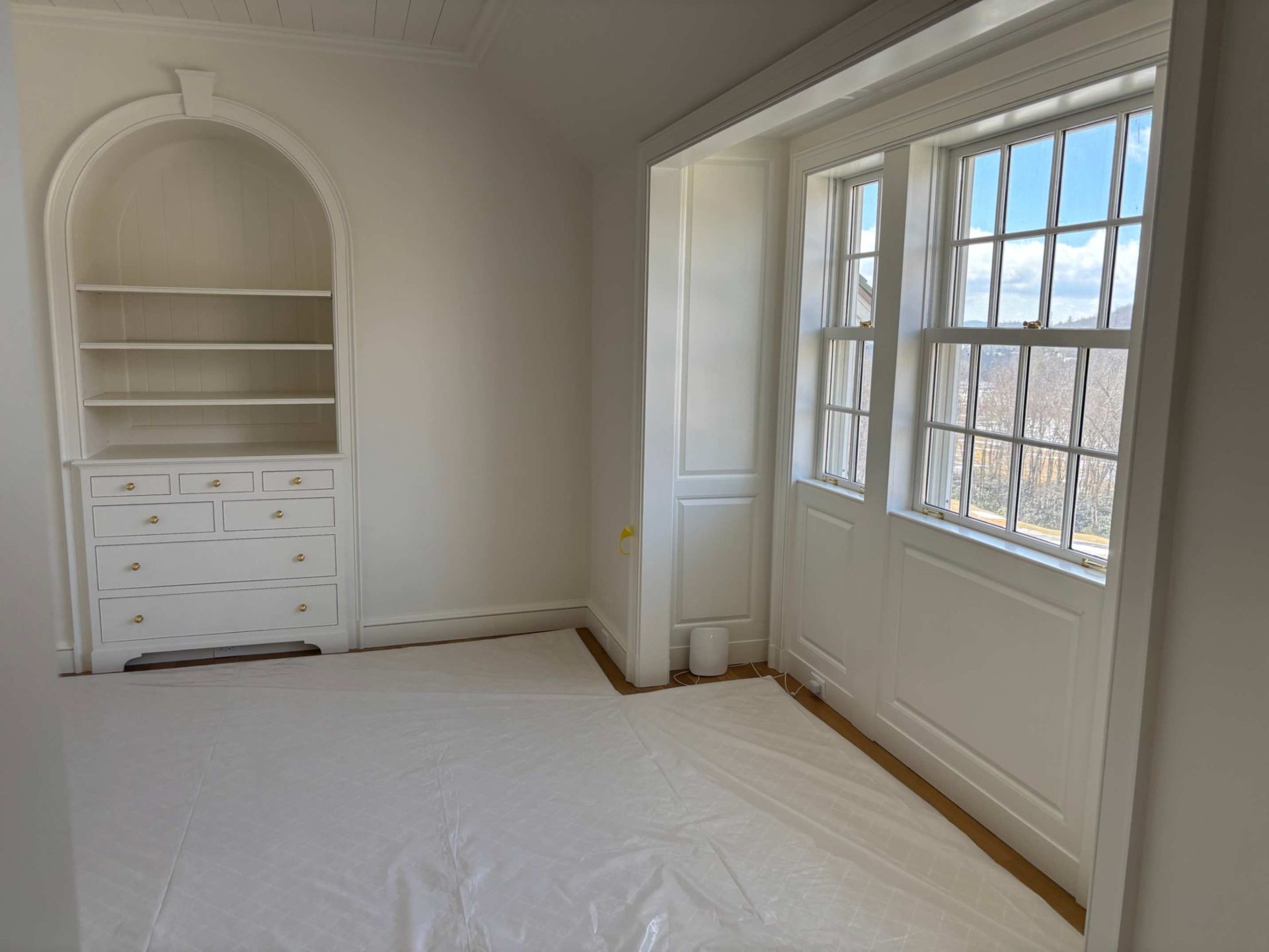 Empty white bedroom with built-in shelves, drawers, and a large window on the right.