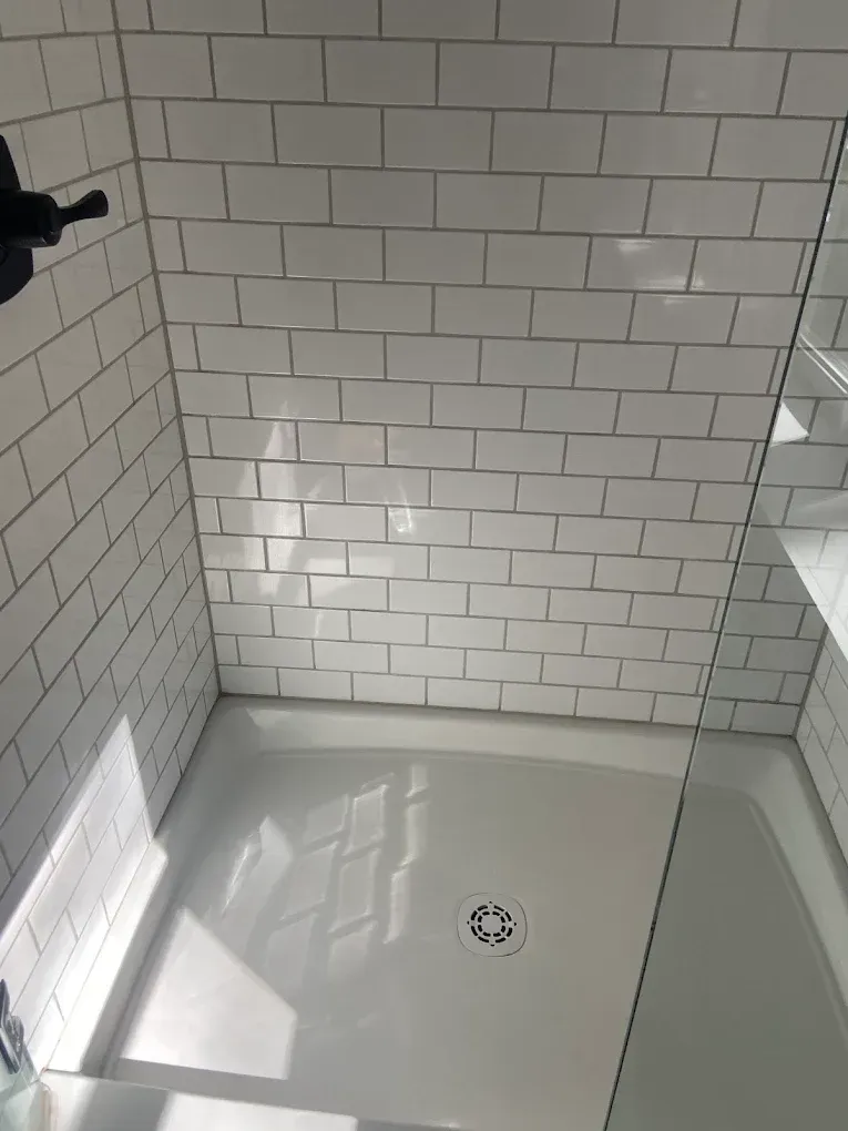 White tiled shower with a flat shower pan and drain, lit by sunlight from a window.