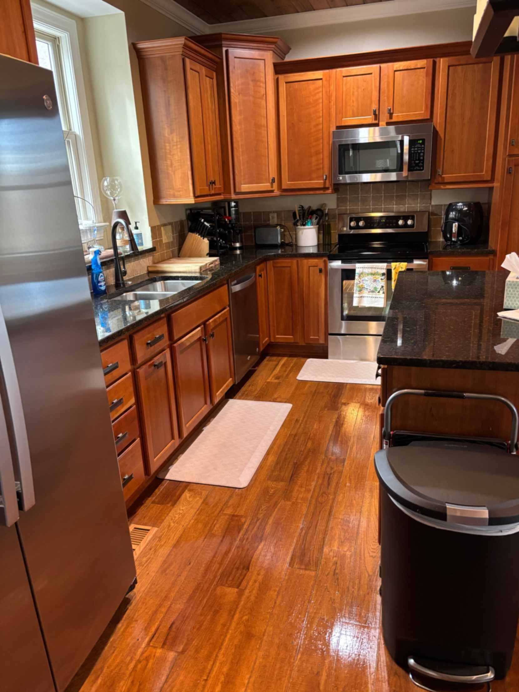 Compact kitchen with wooden cabinets, stainless fridge, black appliances, and a trash can in the foreground.