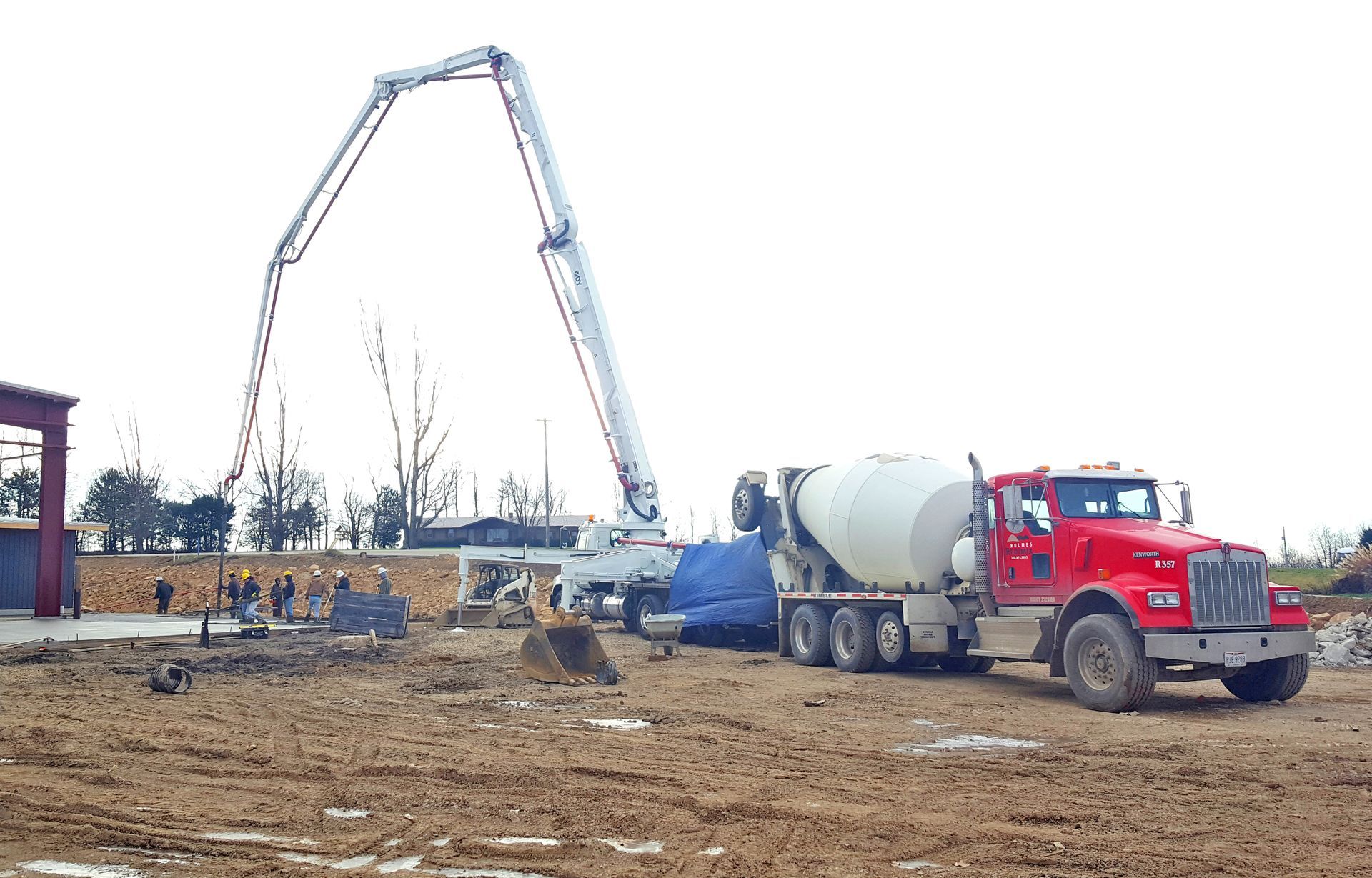 Concrete being poured at a construction site by a boom pump. Red cement truck parked in muddy area.