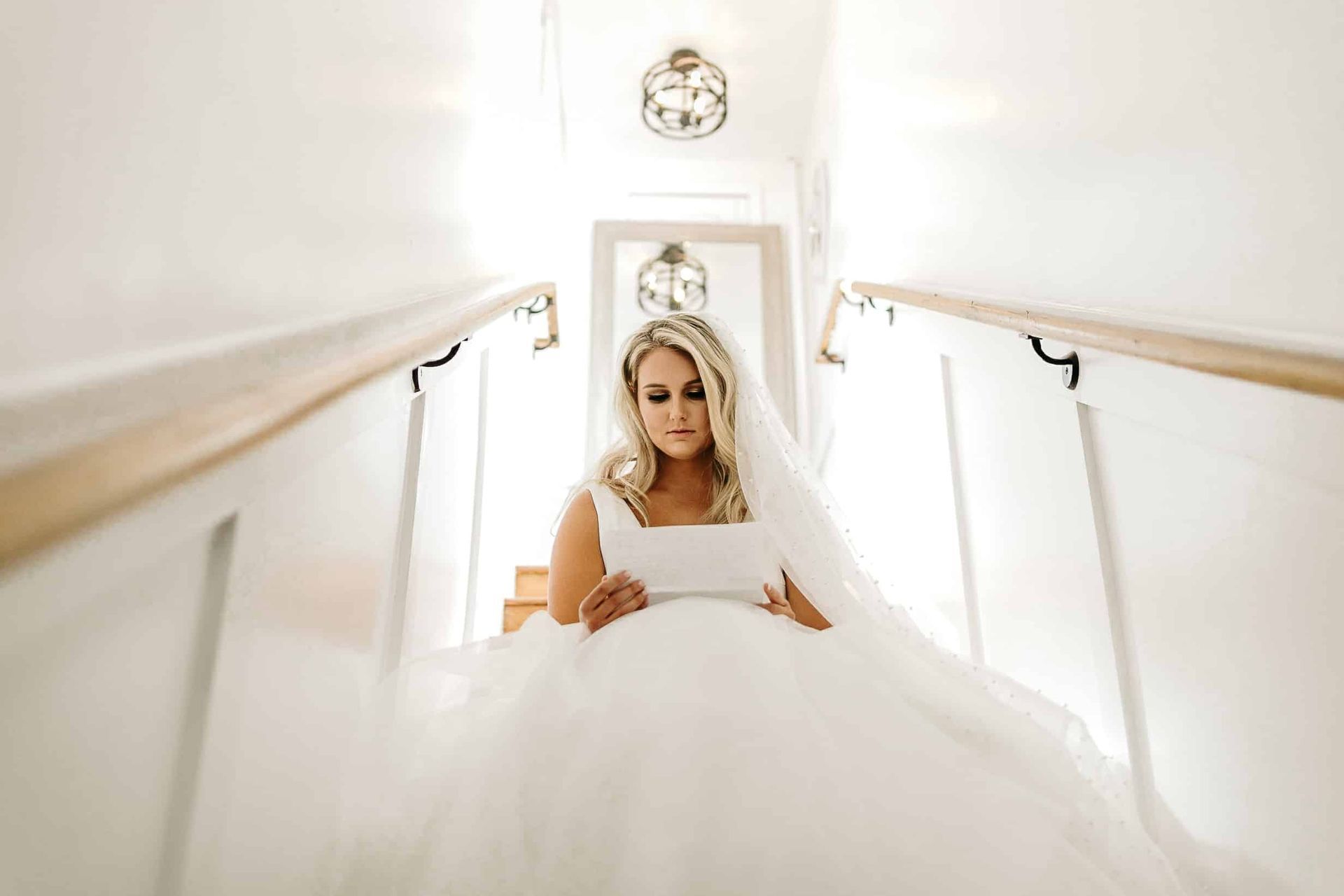 A bride in a wedding dress is sitting on a set of stairs reading a letter.
