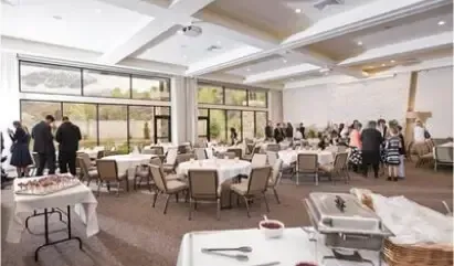 Bright banquet hall with round tables, white chairs, and guests mingling near large windows