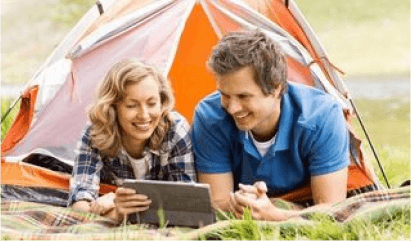 Two people lying in grass outside an orange tent, smiling and using a laptop.