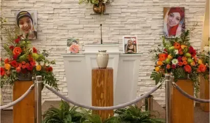 Memorial display with floral arrangements, framed photos, and a white urn behind a rope barrier