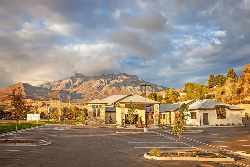 Small building complex with parking lot, trees, and mountains under a cloudy sky