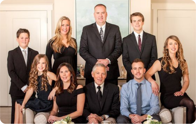 Formal group portrait of eight people posed indoors in front of a bright wall, dressed in business attire