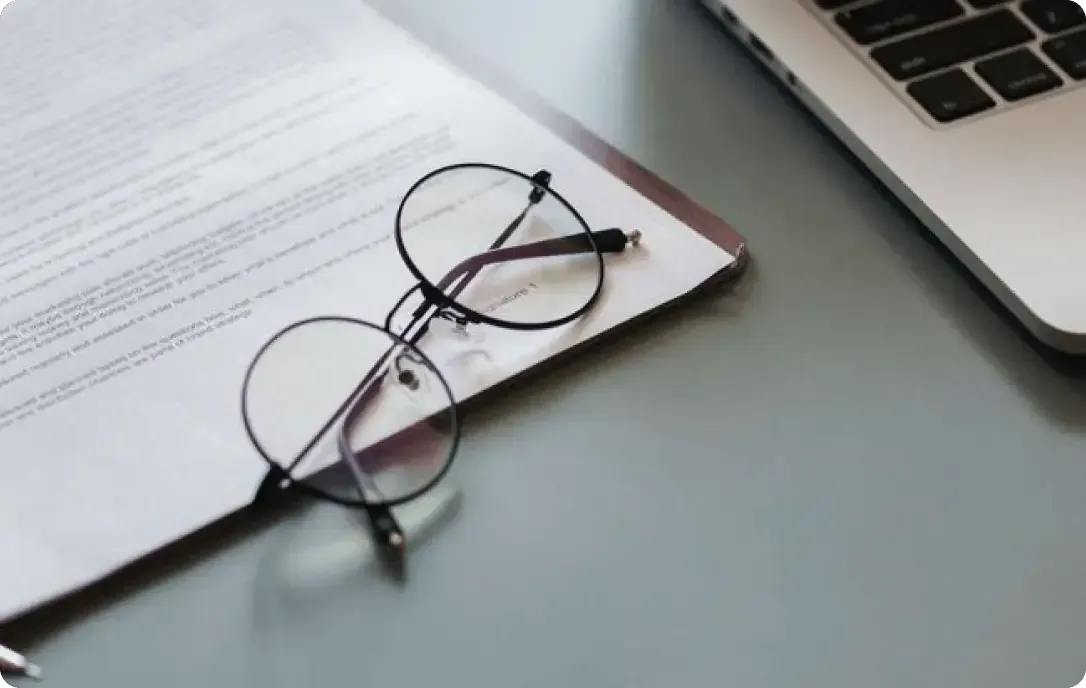 Glasses on an open book beside a laptop on a desk