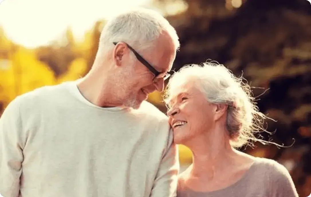 Two smiling people lean close together outdoors in warm sunlight with golden trees in the background