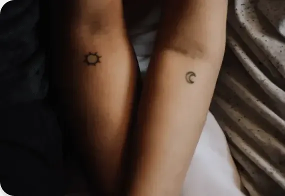Two forearms with small sun and crescent moon tattoos on a bed.