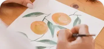 Hands painting orange fruit and green leaves on white paper with a brush