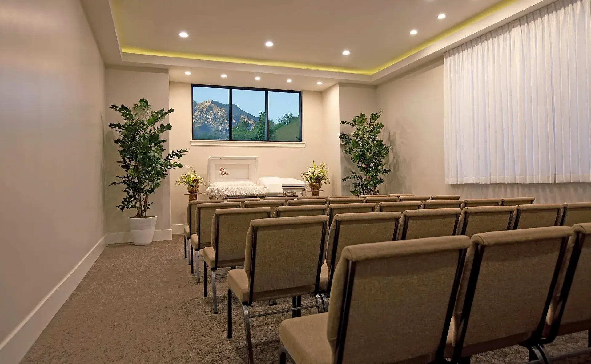 Empty funeral chapel with rows of chairs facing a white casket and floral arrangements under soft ceiling lights