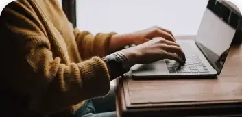 Person typing on a laptop at a wooden desk beside a window