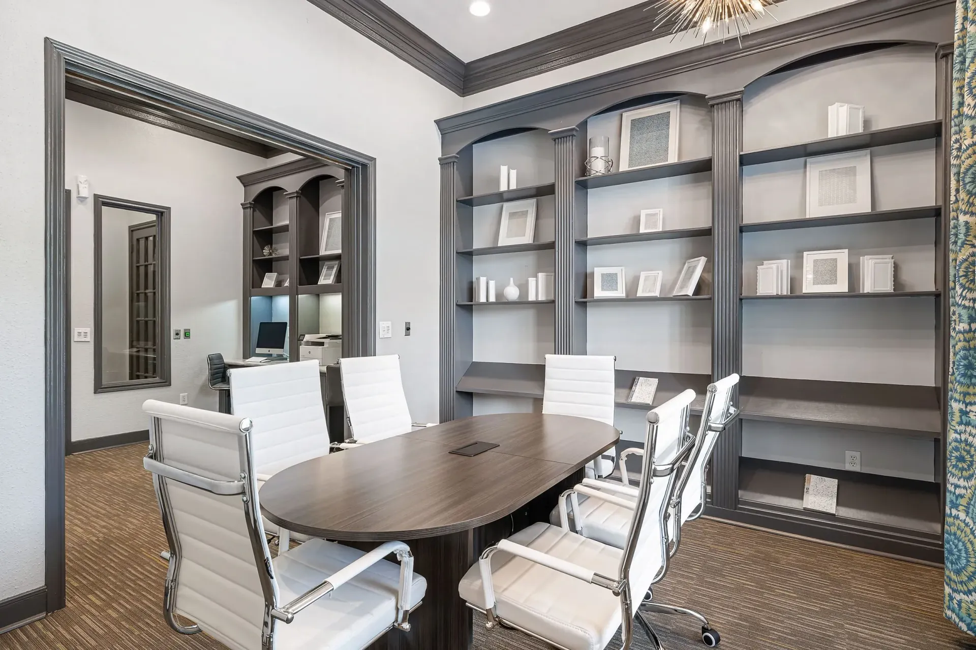 Conference room with oval table and white chairs, bookshelves in background.