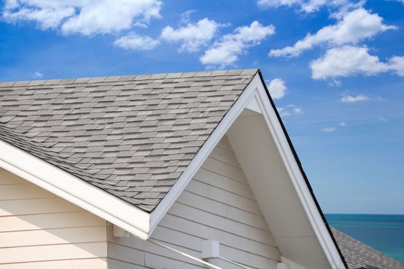 Close-up of a house roof with gray shingles against a blue sky with clouds.