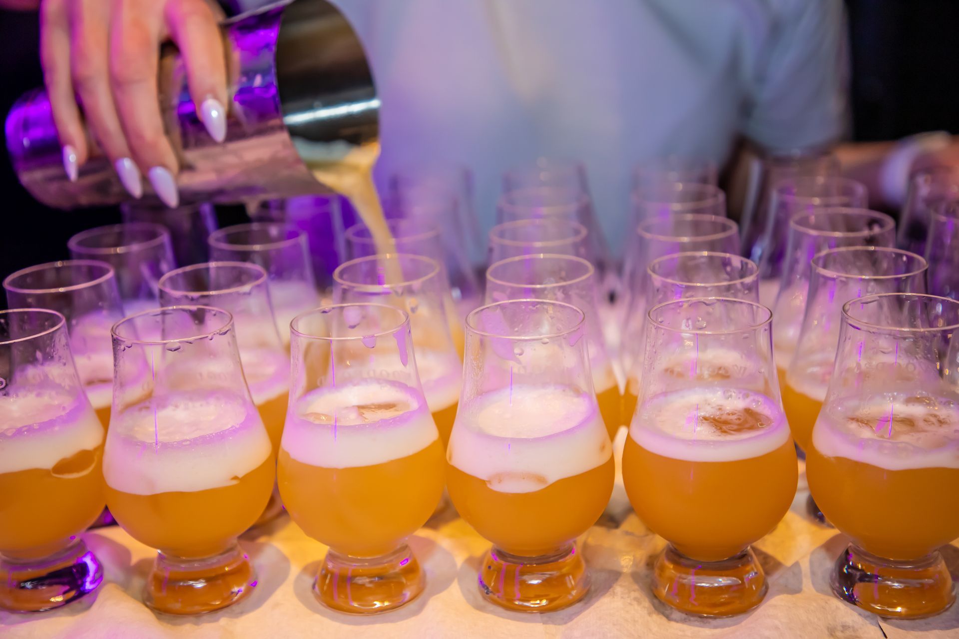 A bartender pours a light orange drink into multiple stemmed glasses in a row, foamy head forming.