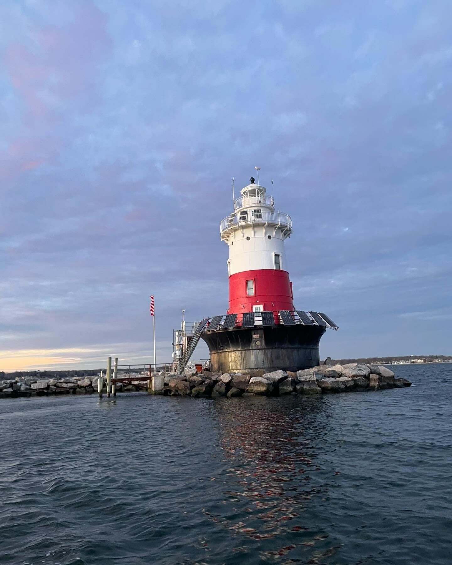 Lighthouse with red and white paint on a rock base in the water under a cloudy sky.