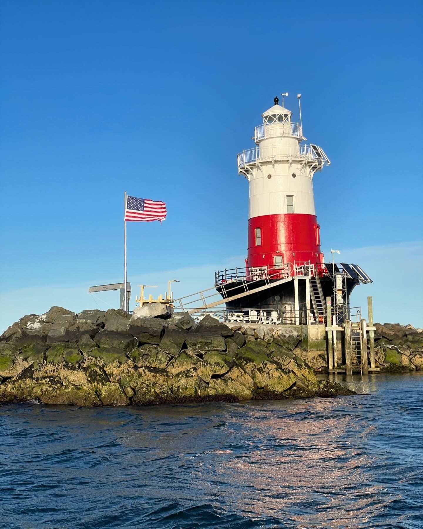 Lighthouse with red and white tower, American flag, on rocky island, blue sky.