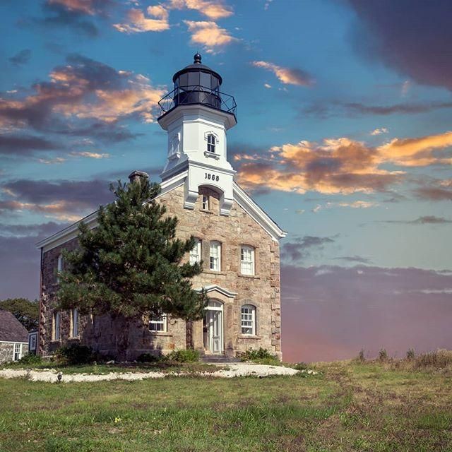 Stone lighthouse with black lantern room against a sunset sky over a grassy field.